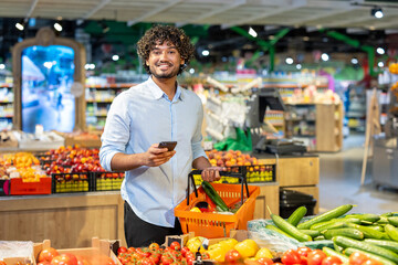A smiling man at the grocery store holds a basket of produce and a smartphone, looking at the...