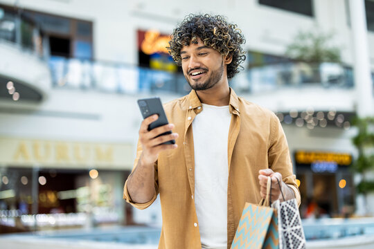 A smiling man looking at his phone while carrying shopping bags inside a modern shopping mall. He seems happy with his purchase.
