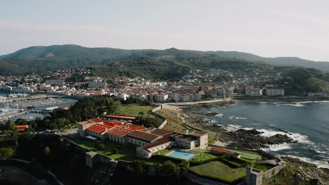 Aerial View Over Parador Hotel On Top Of Oceanside Hill With Baiona Town and Mountains in Background, Spain