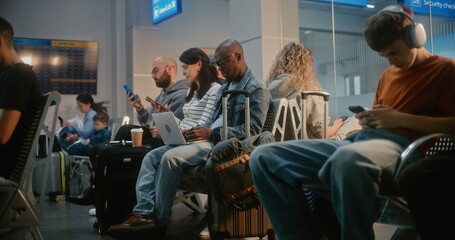 Fototapeta premium Crowded International Airport Terminal: Diverse People, Passengers Sitting in Departure Lounge of Airline Hub, Waiting for Airplane Flight, Using Phones. African American Man Working Online on Laptop.