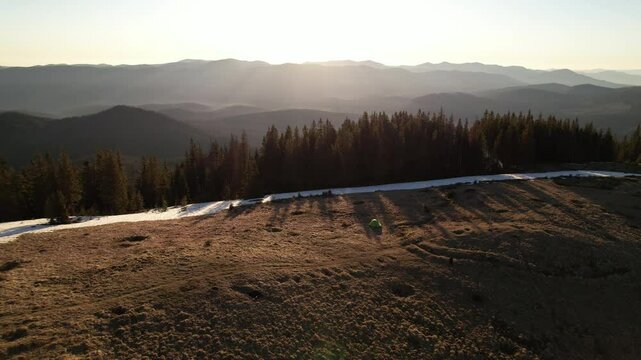 Aerial view of lone green tent on sunlit, grassy plateau near patch of snow, overlooking vast forest and mountain range. Rising sun casts long shadows. Rolling terrain, distant peaks under clear sky.