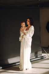 Loving mother in beautiful white dress hugging her naked baby girl while posing in the studio