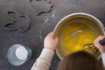 Overhead view of a child mixing yellow batter with a whisk in a metal bowl, surrounded by sugar and...
