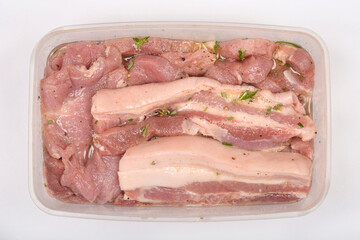 A top-down view shows pieces of raw pork belly marinating in a clear plastic container, seasoned with herbs and spices, against a white background