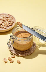 Peanut butter in glass jar on yellow background with knife and salted nuts.