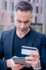 Confident caucasian man with headphones serious while using tablet and holding a credit card outdoors. Concept of online shopping, digital payment, modern lifestyle, and financial technology. Vertical
