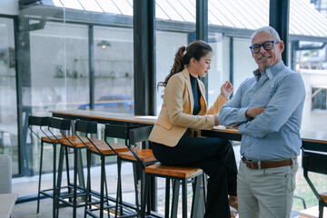 Senior businessman with crossed arms listening to young businesswoman explaining something during informal meeting at modern office cafeteria
