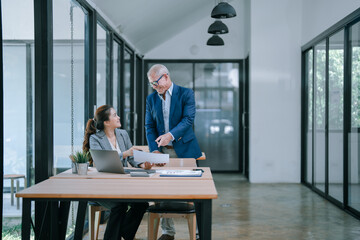 Senior manager explaining paperwork while seated at a desk, engaging with a young businesswoman using a laptop in a modern office with glass walls