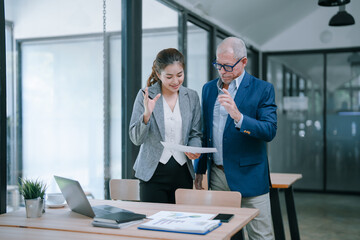 Senior businessman and young businesswoman are standing near a desk and discussing and analyzing a financial document together in a modern office, collaborating and working together as a team