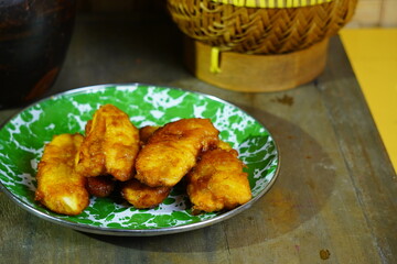 A still life of Indonesian food featuring fried bananas on a plate, a brown clay jug, a woven bamboo basket, and fresh bananas on a wooden surface with bamboo backdrop.