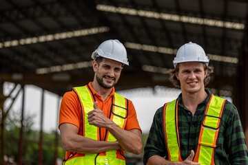 Two male civil engineers express strong confident with thumb up in front of a construction site. Partnership between two supervisors or factory managers. Workers in safety suits and helmets.