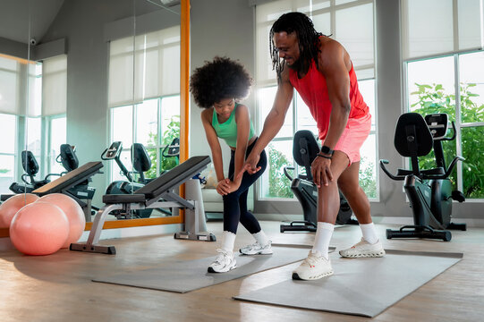Black father is fitness trainer supports his daughter stretching her leg on a yoga mat in a modern gym filled with exercise equipment.
