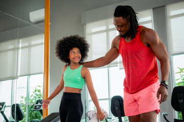 Black father as a fitness trainer helps his daughter stretch and balance in a modern gym filled...