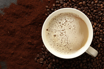Cappuccino, top view. Cup of cappuccino among coffee beans and ground coffee. Cup of aromatic coffee among roasted coffee beans. Invigorating drink. Copy space