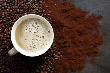 Cappuccino, top view. Cup of cappuccino among coffee beans and ground coffee. Cup of aromatic coffee among roasted coffee beans. Invigorating drink. Copy space