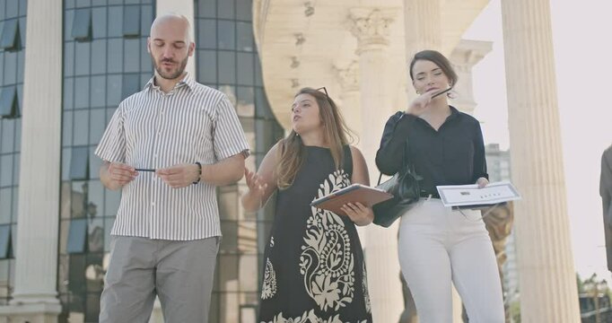 Business colleagues walk and talk outside, discussing a project on a sunny summer day near classical statues and columns.