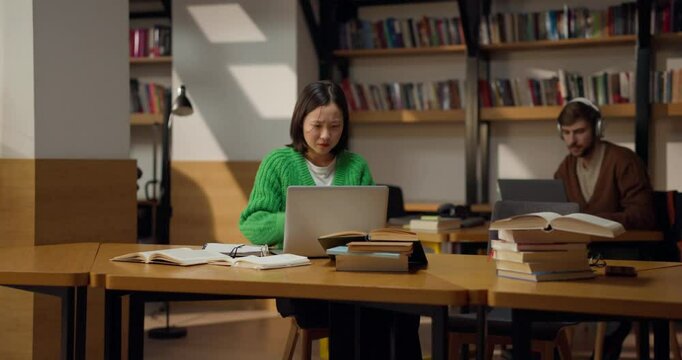 Female student in a green sweater studies at a laptop in a cozy library surrounded by books and notes