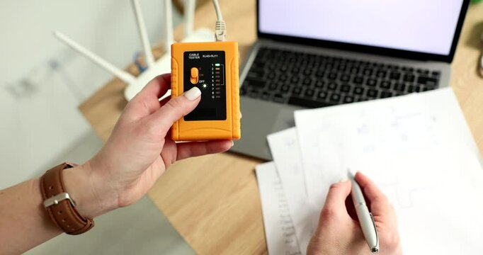 Female network technician checks Internet cable using tester and documenting findings. Laptop and router at desk during network maintenance check