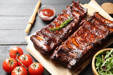 Tasty roasted pork ribs served with rosemary, tomatoes, sauce and salad on black wooden table, closeup