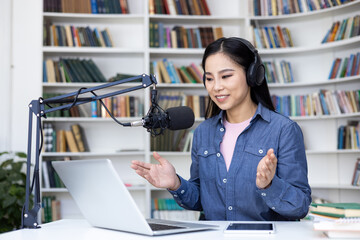 An asian woman records a podcast in front of a bookcase. She wears headphones and talks into a microphone with a laptop open.