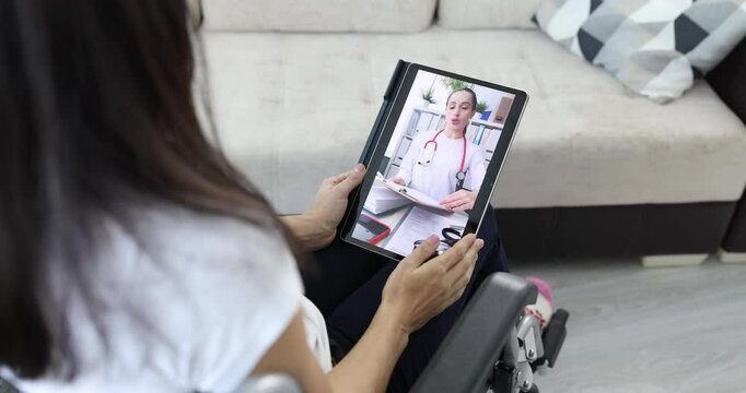 Female patient with disability sitting in wheelchair holds tablet during video call appointment with doctor woman. Online medical consultation at home
