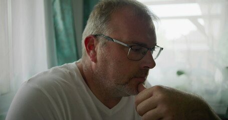 Man gazing pensively while resting his chin on his hand, seated near a window with soft green...