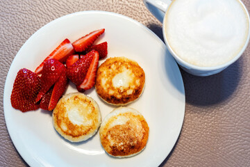 A delicious and healthy breakfast of syrniki served with fresh strawberries and a cup of frothy cappuccino. Traditional Eastern European dish