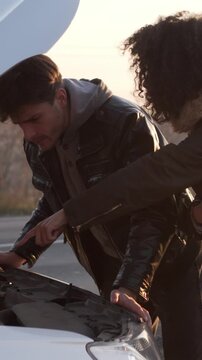 Concerned wife and husband examines broken car with the open hood on the road outdoors, facing vehicle troubles during trip. Vertical video.