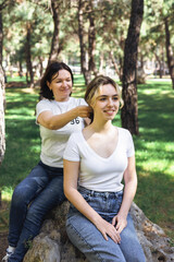 Fototapeta premium Smiling mother tying teenage daughters hair while sitting outdoors