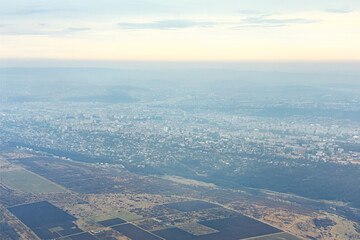 Aerial view of Chisinau cityscape with numerous buildings spread across vast area, surrounded by patches of greenery and agricultural fields. Overcast sky and city appears is enveloped in haze