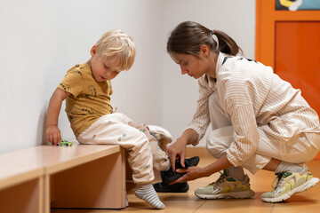 A mother assists her young child with putting on shoes as the child sits on a bench in a kindergarten or daycare hallway. Focus on daily routines, parental care, and getting ready.