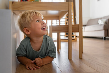 A happy, blonde toddler lies on a wooden floor, smiling broadly and looking up playfully from under a wooden chair. Captures a joyful childhood moment of exploration and fun in a bright room.