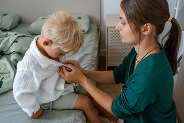 A mother gently helps her blonde toddler son get dressed, buttoning his white shirt as he sits on...
