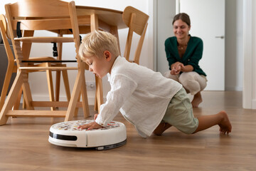 A young blonde child curiously interacts with a robotic vacuum cleaner on the wooden floor at home....