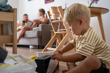 A young child sits on the floor, focused on playing with toy trucks, while parents relax on the sofa in the blurred background. Scene capturing independent playtime within a family home.