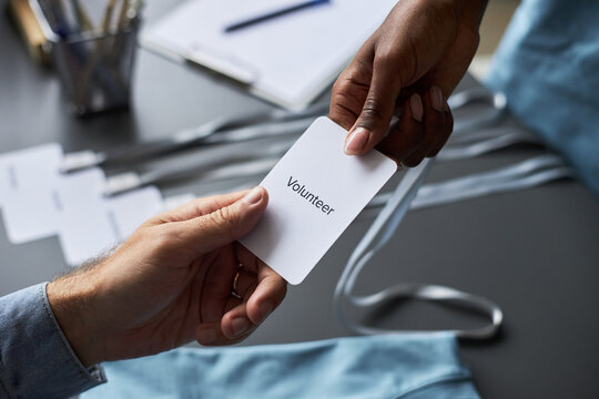 Close-up of person handing a volunteer badge to another person at event registration desk. Badges and lanyards are visible on table, suggesting an organized event environment