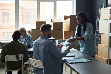 Team of diverse individuals collaborating on packing clothes for shipping in well-lit room with large windows and stacked boxes. Elderly man and young adults involved in project