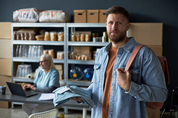 Portrait of man standing and holding folded clothes in warehouse with wooden shelves filled with...