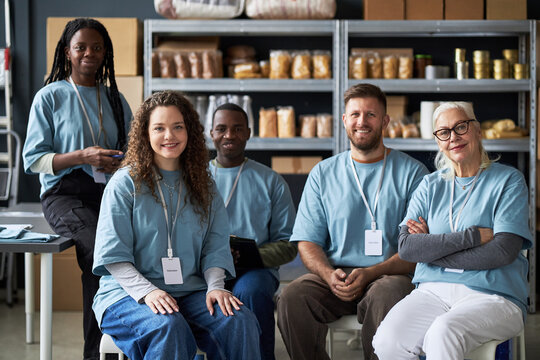 Portrait of diverse group of volunteers posing in community center with smiling expressions, wearing matching shirts and name tags while sitting in front of shelves full of supplies