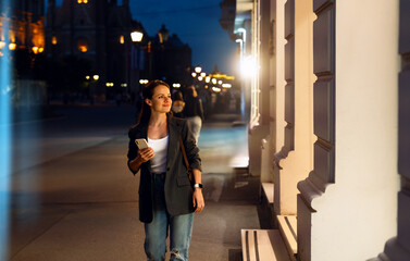 Young woman walking at night after work and looking at illuminated shop window