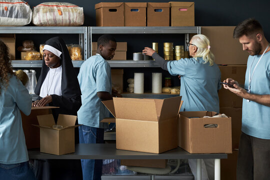 Diverse group of volunteers organizing food items in boxes for community donations Standing in storage area surrounded by shelves filled with supplies