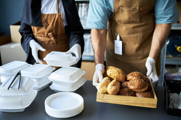 Plakat Restaurant staff preparing food orders with hands seen organizing takeaway boxes and serving freshly baked bread on a tray in the kitchen. Workers wearing aprons and gloves