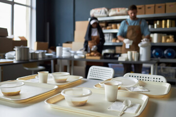 Volunteers preparing meals in a community kitchen, carefully arranging trays with bowls, cups, and cutlery. Shelves filled with supplies in background emphasizing activity and teamwork