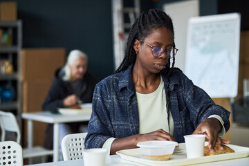 Woman wearing eyeglasses and cardigan eating lunch in a room with tabletop and chairs. Senior...
