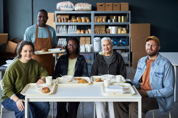 Group of people from diverse backgrounds enjoying meal together at a community center with trays of food and drinks on table. Shelves with bread and other items in background