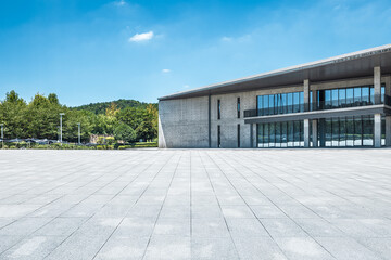 Empty stone square floor in front of a modern building exterior with green trees under a blue sky.