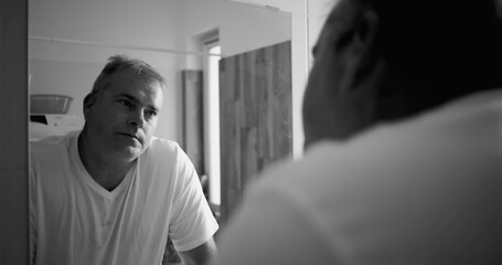 Man reflecting in bathroom mirror, black and white tone, thoughtful expression, hands on counter, wooden bathroom details in background, calm and serene atmosphere