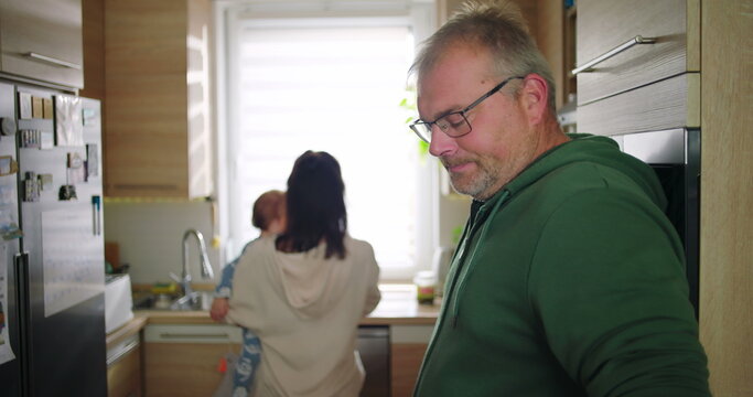 Man smiling with cup in hand while mother holds baby in kitchen, cheerful family moment during everyday home routine, casual and warm interaction