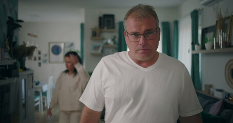 Man staring at camera with intense expression during argument, woman in background in living room, tense and emotional moment of relationship conflict and frustration