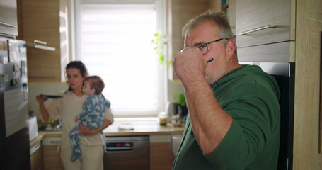 Man stirring drink in kitchen while mother holds baby in background, family multitasking during morning routine, everyday domestic life at home